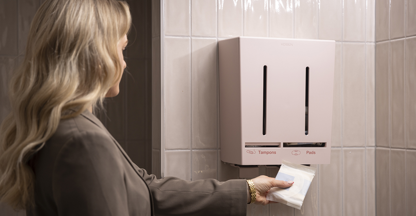 Kvinna tar ett mensskydd från Herbox rosa dispenser i ett kontorstoalettutrymme.
