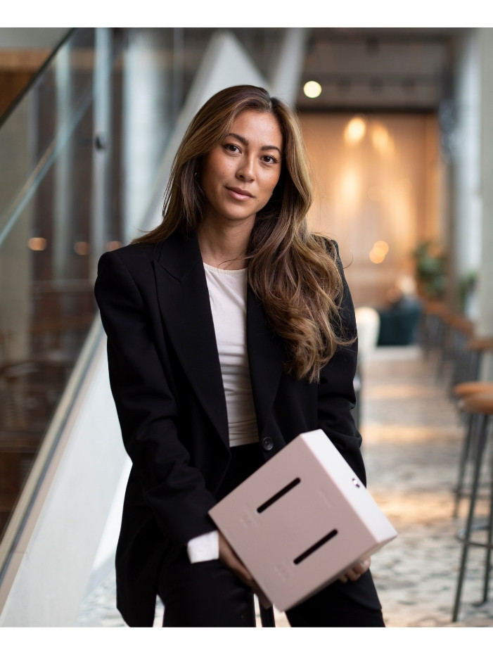 Hanna Lauste i svart kavaj håller en rosa herbox inomhus, med suddig bakgrund.