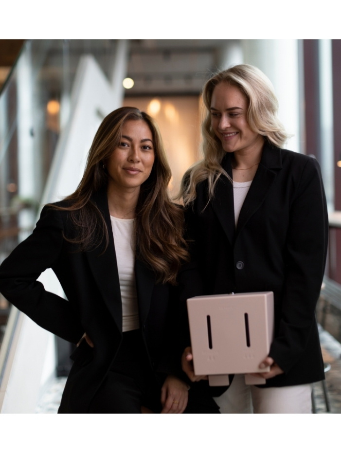 Hanna Lauste och Linn Rosenqvist i svarta kavajer, Linn håller en rosa automat för mensskydd, står inomhus och ler.