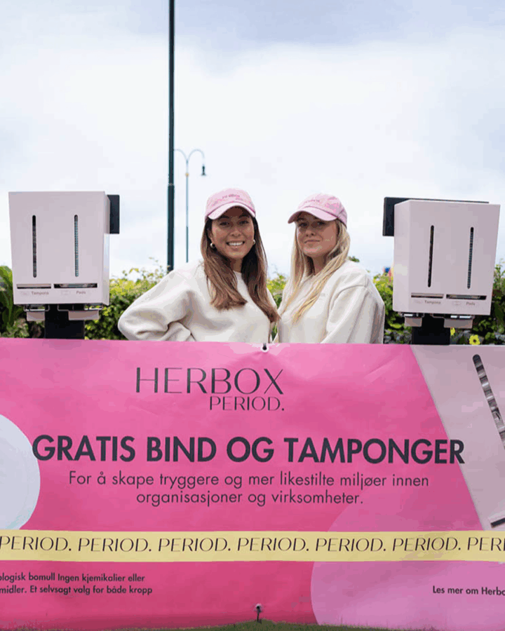 Hanna och Linn i rosa kepsar står framför en skylt om gratis bindor och tamponger från Herbox.