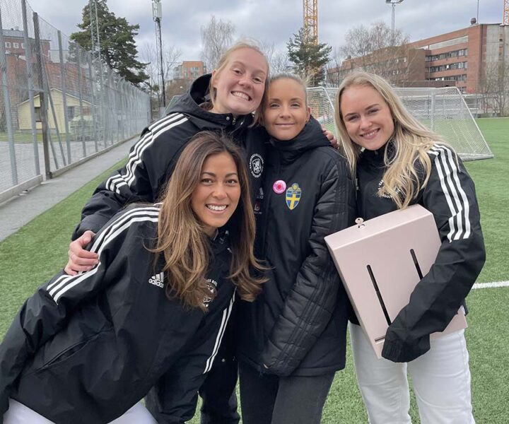 Hanna och Linn från Herbox står tillsammans med Svenska Fotbollförbundet och en rosa mensskyddsautomat på en fotbollsplan, ett initiativ för att tillgängliggöra gratis mensskydd inom idrotten