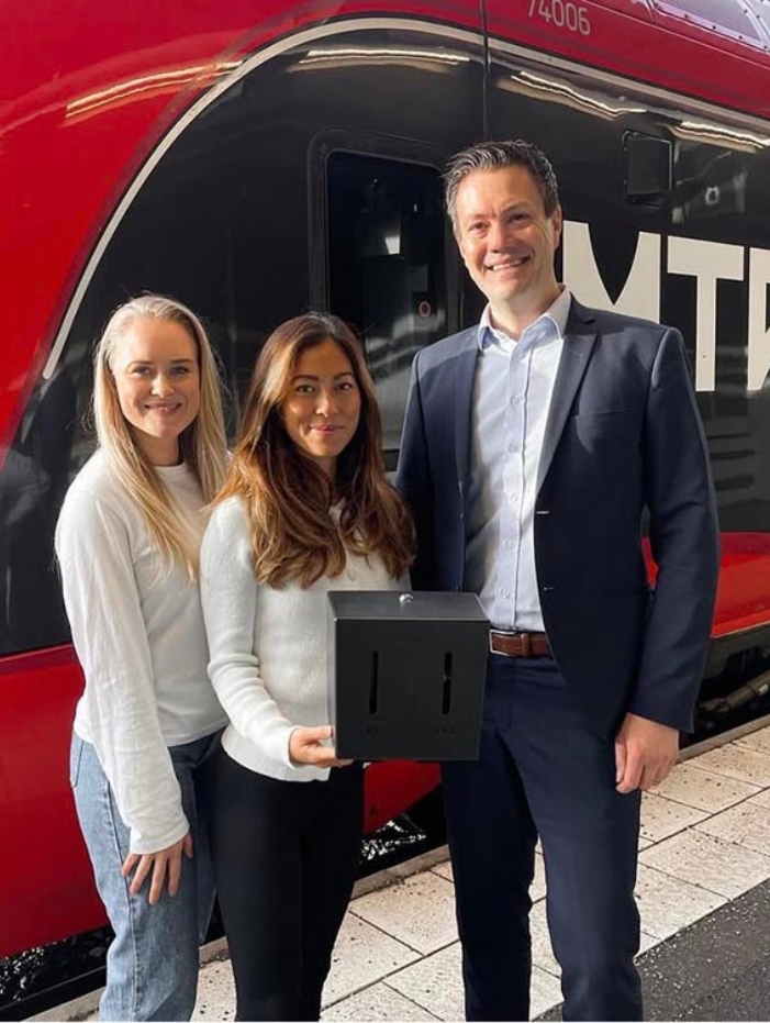 Bild på Linn Rosenqvist och Hanna Lauste grundare av Herbox tillsammans med en representant från MTRX (VR tåg) som visar upp automaterna som finns ombord på alla deras tåg. Med ett rött MTRX tåg i bakgrunden.