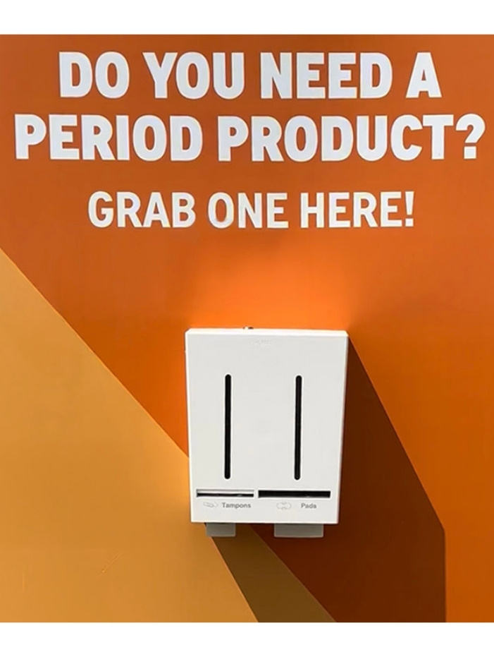Vit behållare för tamponger och bindor från Herbox på orange vägg, med texten "DO YOU NEED A PERIOD PRODUCT? GRAB ONE HERE!". På Roskilde festival
