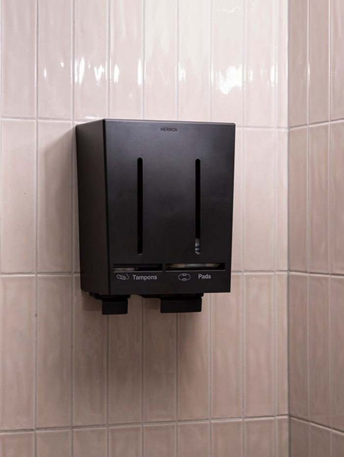 Svart Herbox dispenser för tamponger och bindor monterad på en ljust kaklad vägg.