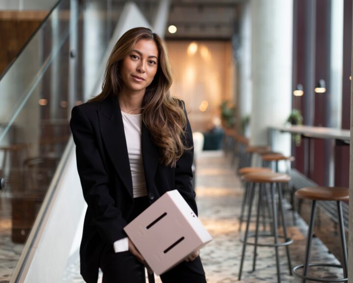 Hanna Lauste i svart kavaj håller en dispenser inomhus i en stilren miljö med barstolar och fönster.