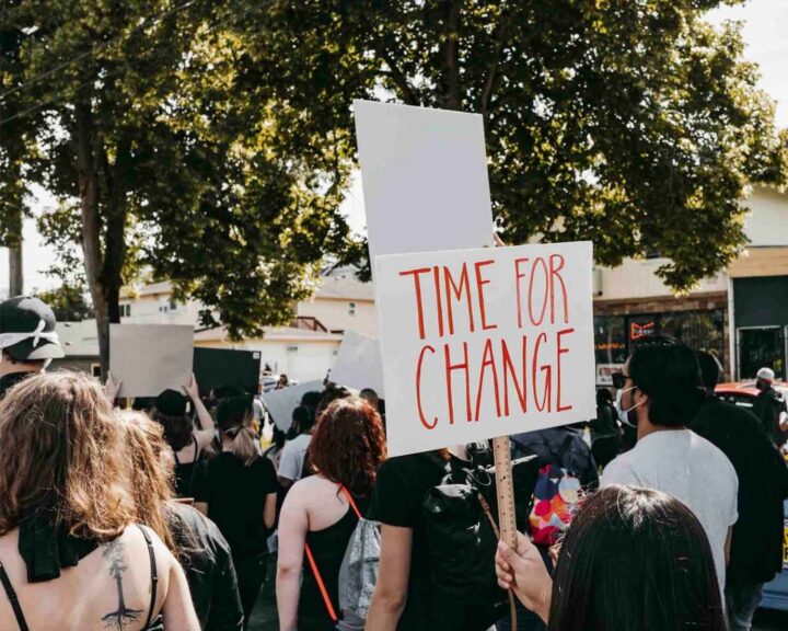 Protest med folk som håller upp skyltar. En skylt lyder "Time for Change". Träd och byggnader i bakgrunden.