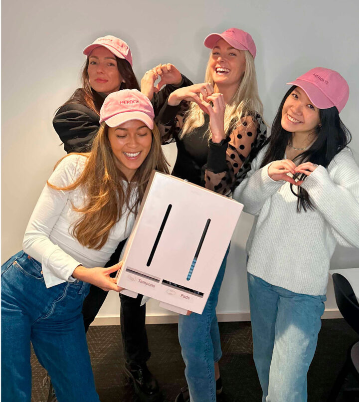 Fyra kollegor från Herbox med rosa Herbox kepsar poserar glatt med en låda för mensskydd.