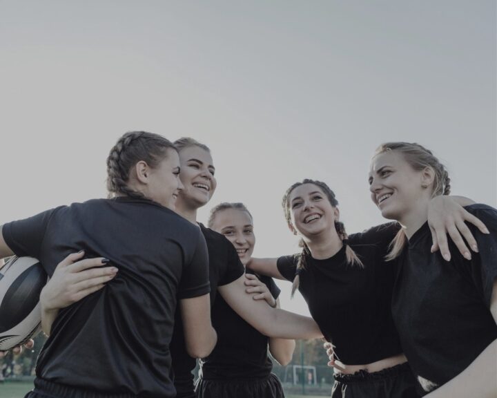 Fem kvinnor i svarta sportkläder kramas och skrattar utomhus.