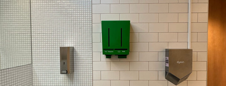 På en vägg hänger en tvålbehållare, en grön tampong- och bindaautomat från Herbox och en Dyson handtork.