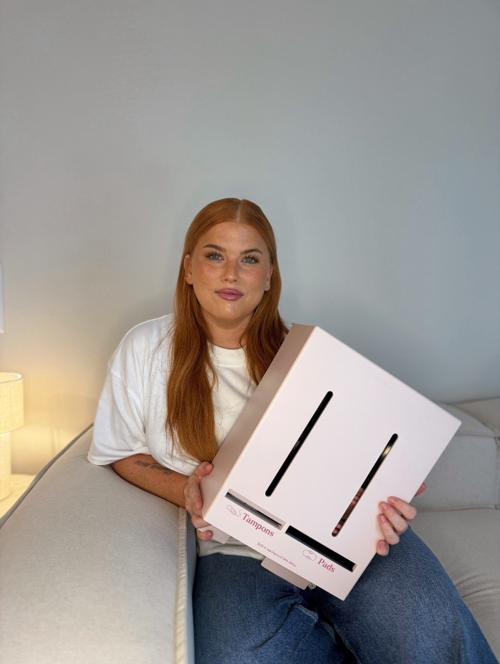 Jessica "Mithanja" med långt rött hår sitter på en soffa och håller en rosa låda för tamponger och bindor från Herbox.