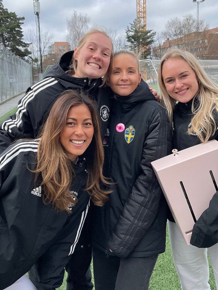 Fyra kvinnor i sportjackor står nära varandra och ler på en fotbollsplan. Herbox + SvFF = sant