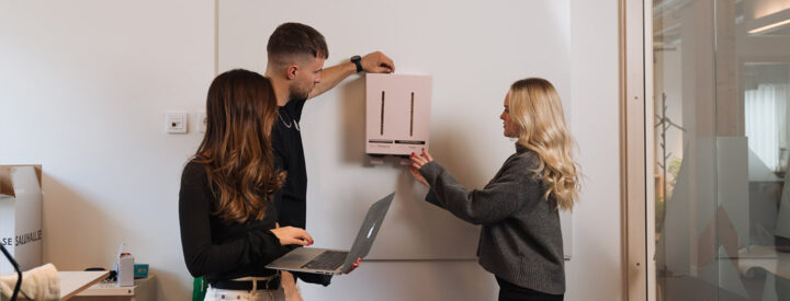 Tre personer installerar en rosa dispenser på en vägg från Herbox. En person håller en laptop.