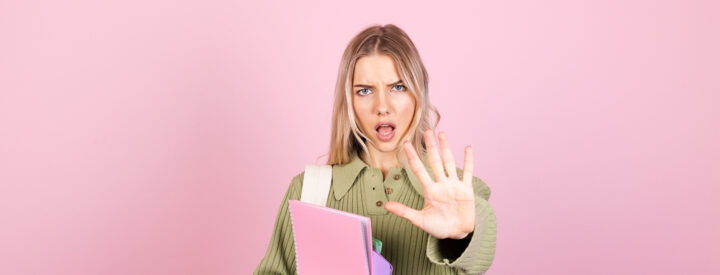 Kvinna med grön tröja och block, håller upp handen mot kameran, rosa bakgrund.