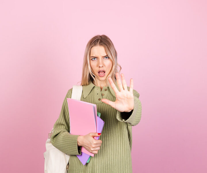 Kvinna i grön tröja framför rosa bakgrund, håller mappar och visar stopptecken med handen.